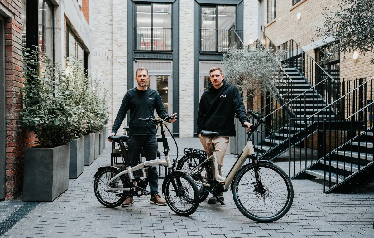 Deux hommes posent dans une cour pavée, chacun à côté d'un vélo électrique VOLT à cadre clair