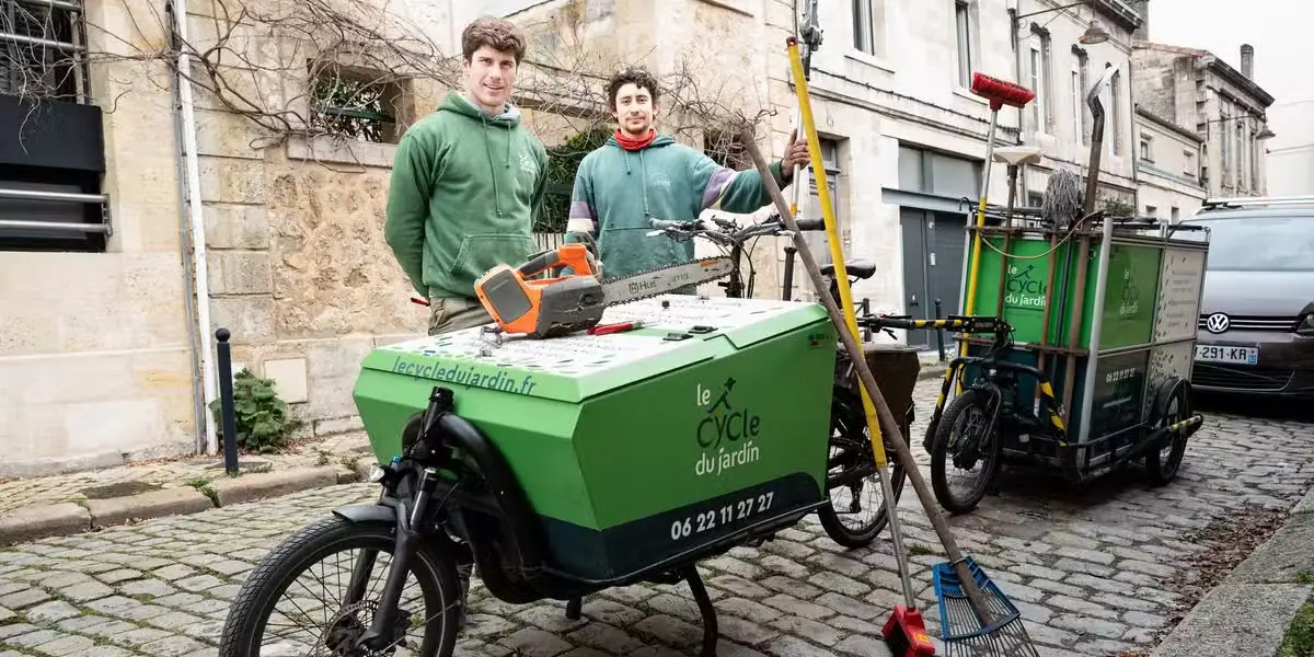 Deux paysagistes debout derrière un biporteur électrique chargé d'outils, tronçonneuse, perches et râteaux, dans une rue pavée