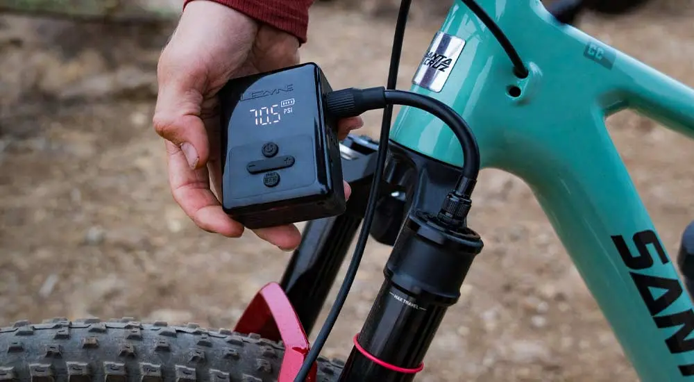Le gonfleur électrique Lezyne E-Drive en main devant un VTT, l'écran affiche la pression en PSI