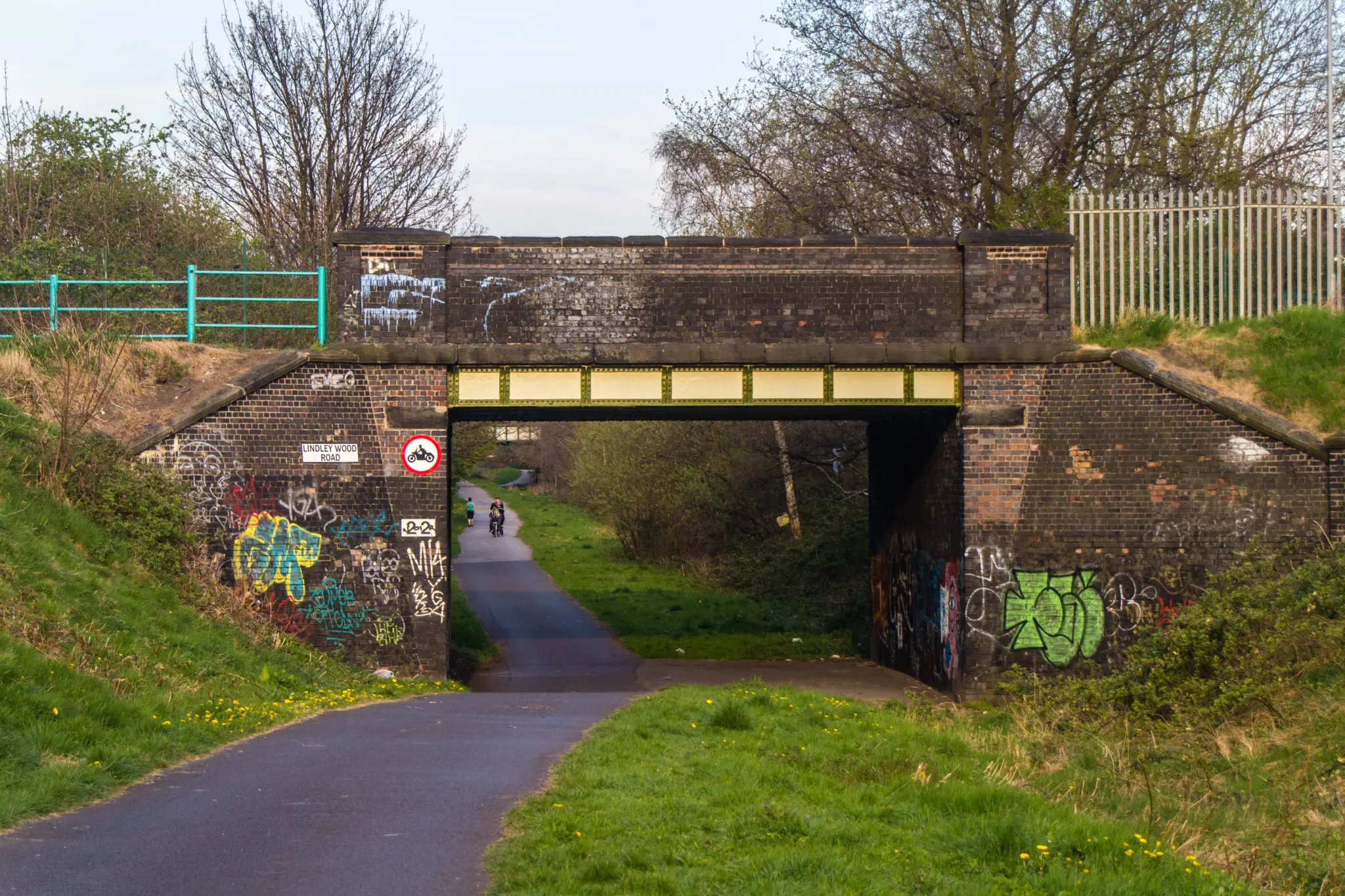 Piste cyclable goudronnée passant sous un pont de brique, au milieu de la végétation, avec un cycliste au loin
