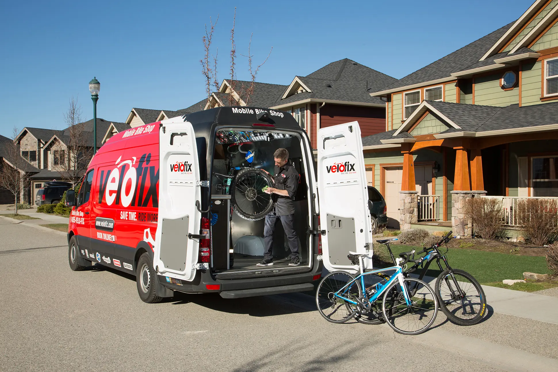 Mécanicien Velofix, racheté par Backcountry, travaillant sur un vélo à l'arrière d'un van ouvert, deux vélos posés sur le trottoir