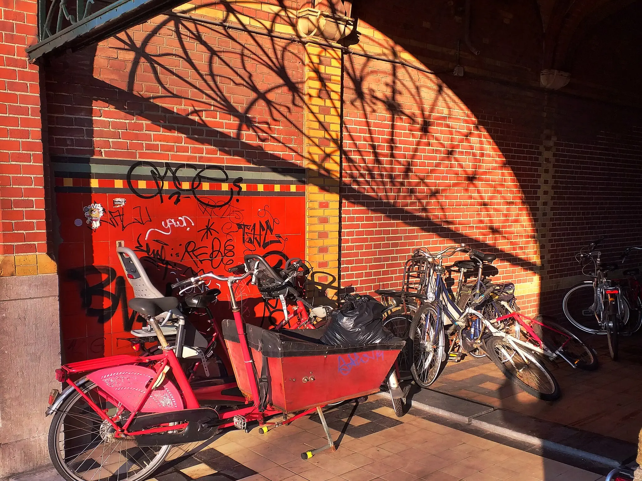 Un vélo cargo rouge équipé d'un siège enfant, garé avec d'autres vélos dans un passage en briques
