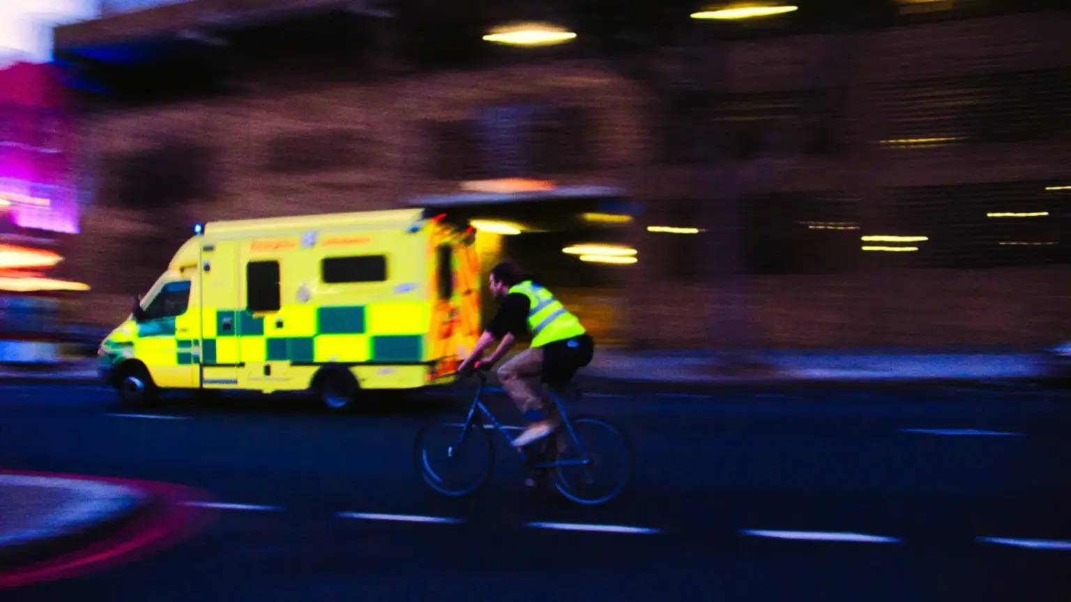 Un cycliste sans casque, en gilet jaune, roule de nuit dans une rue urbaine tandis qu une ambulance passe en arriere-plan