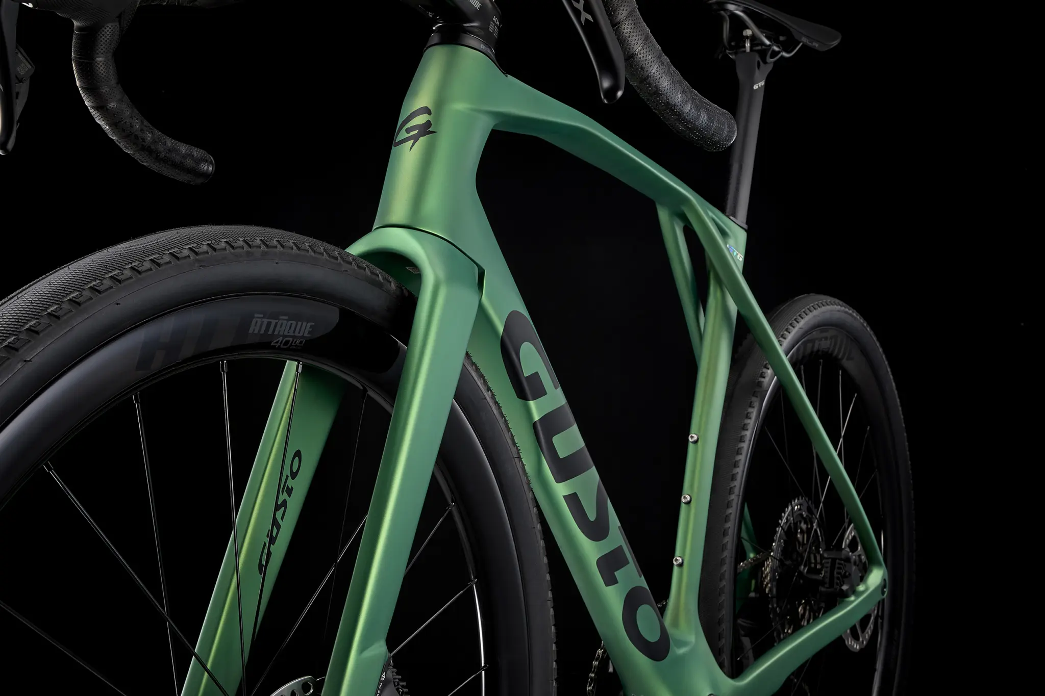 Front of a green Gusto GTG gravel bike, with the Gusto logo on the down tube and a knobbly tyre on a carbon wheel