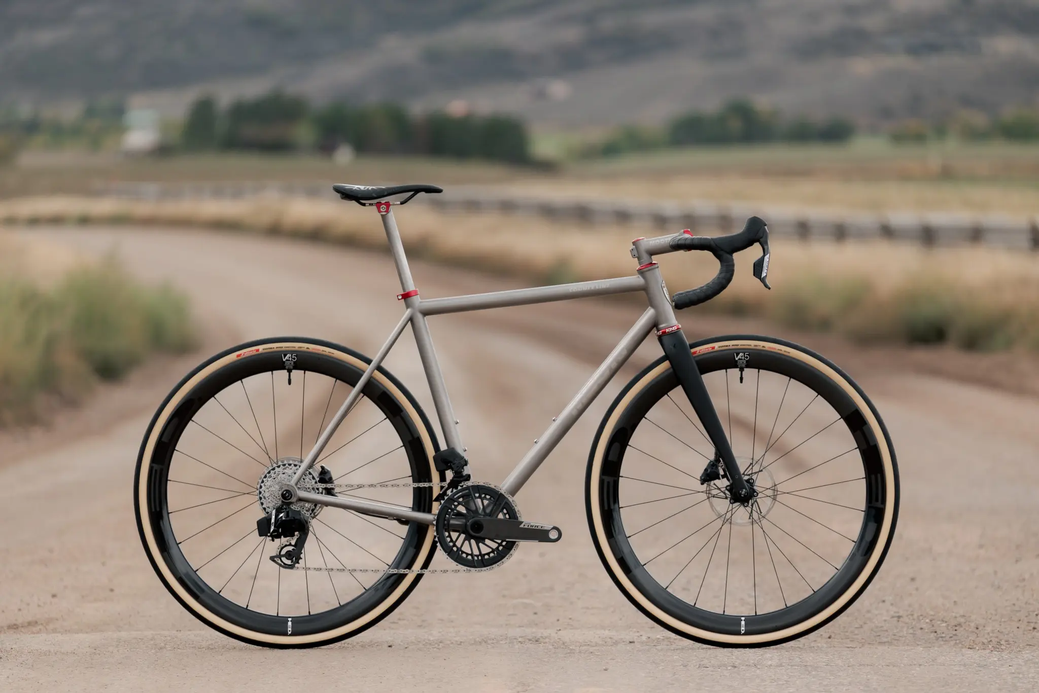 Moots County Line en titane brut de profil sur un chemin de terre, fourche et cintre noirs, roues carbone