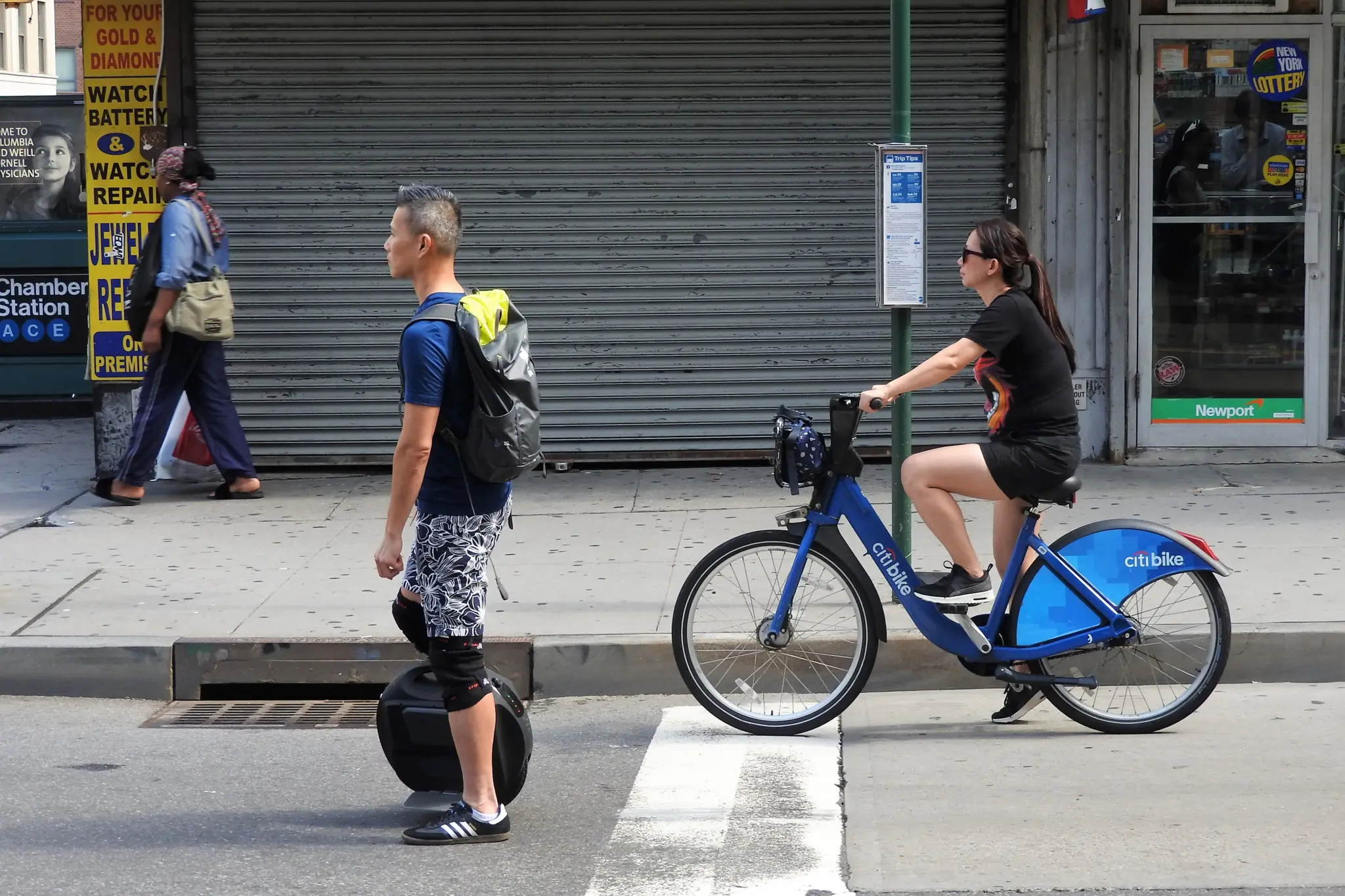 Une cycliste sur un Citi Bike bleu croise un homme debout sur une gyroroue électrique, dans une rue de New York