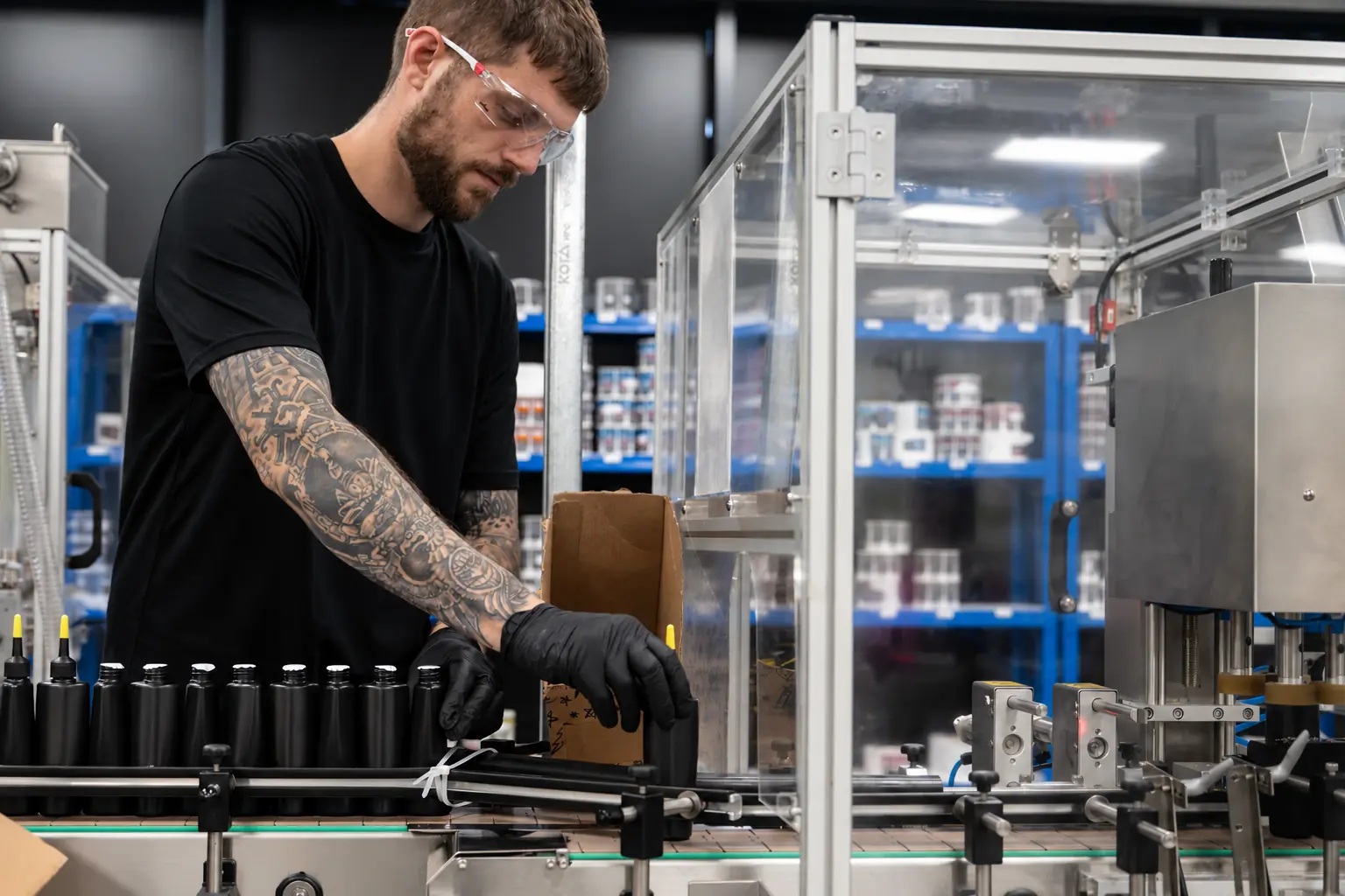 Un opérateur en t-shirt noir, lunettes de protection et gants, place des flacons noirs sur une ligne de conditionnement automatisée (visuel du communiqué Muc-Off)
