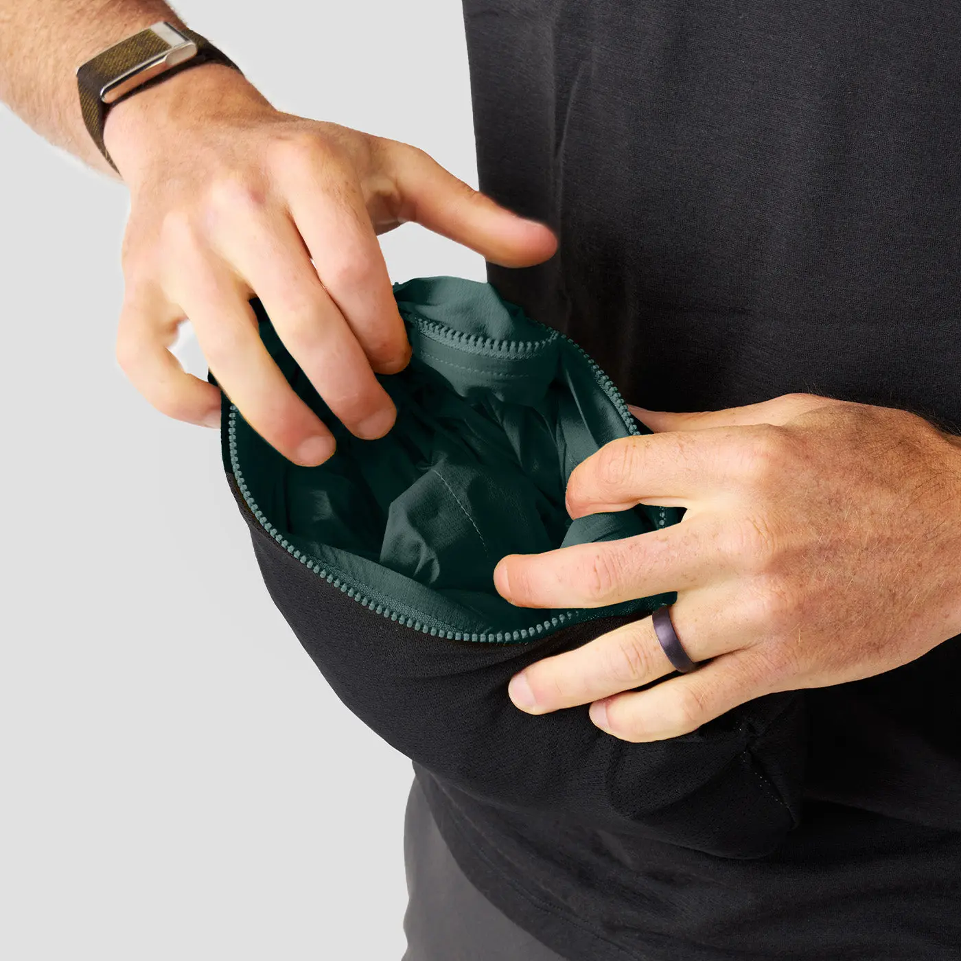 Hands packing the dark green Ornot Fog Eater jacket into its own zipped pocket
