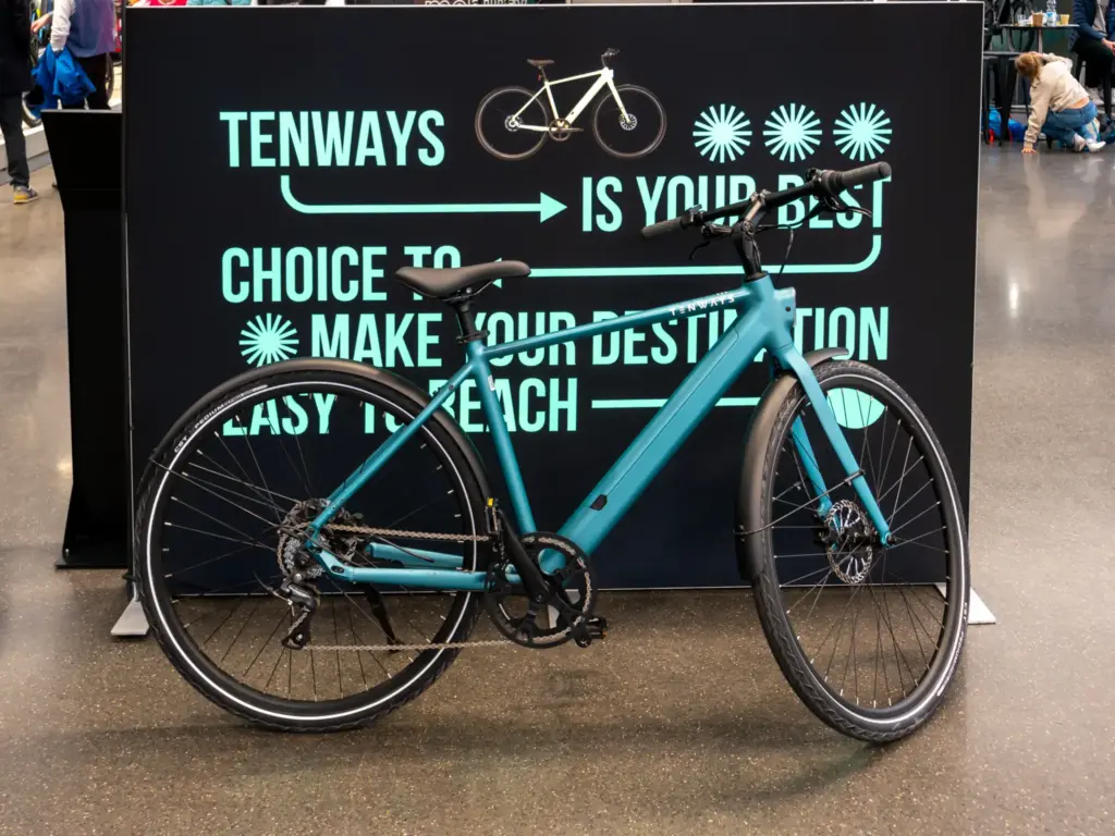 Vélo électrique Tenways bleu-vert devant un panneau noir de la marque, sur un salon