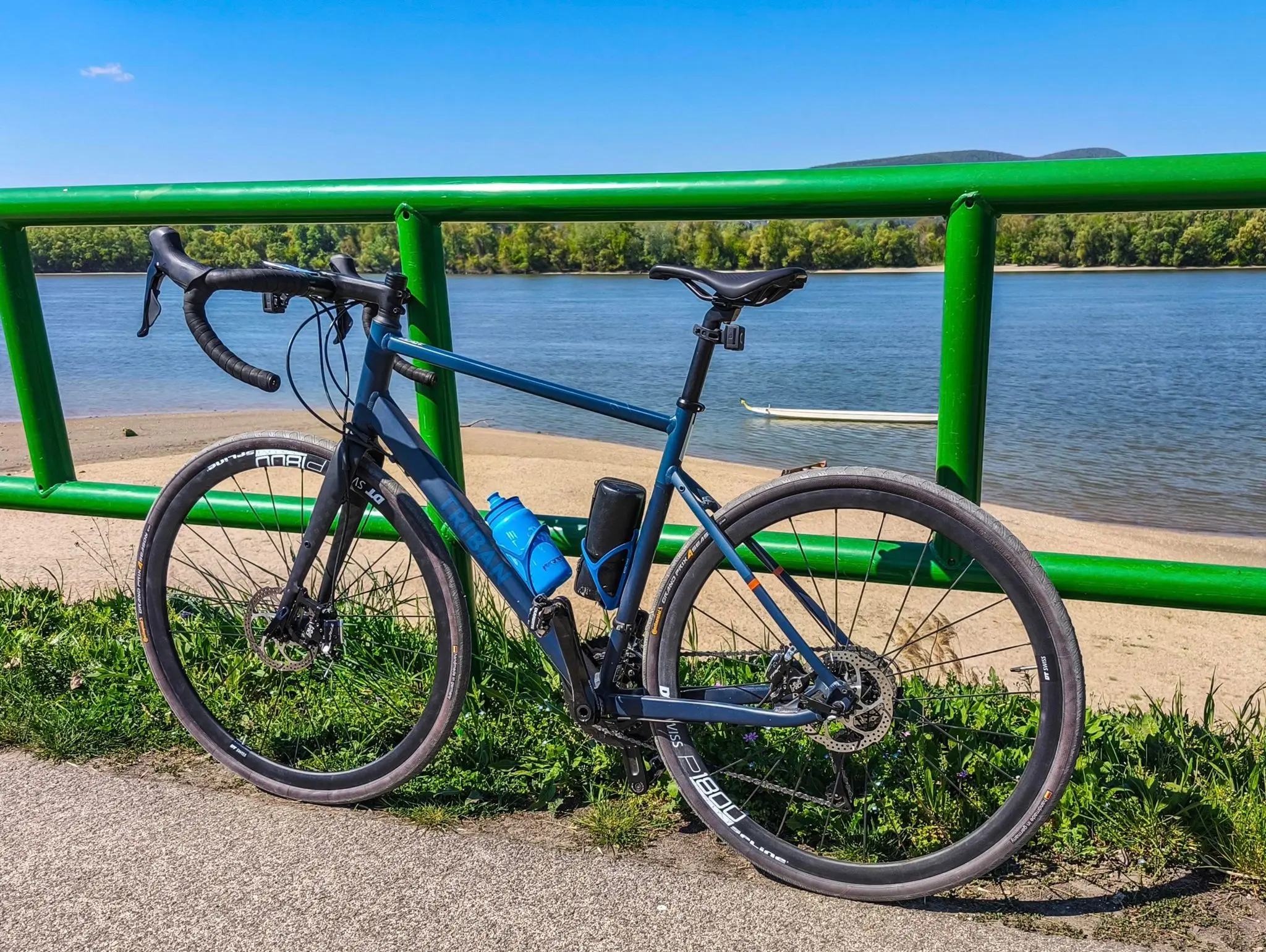 Un vélo de route Triban bleu de Decathlon appuyé contre une rambarde verte au bord d'un fleuve