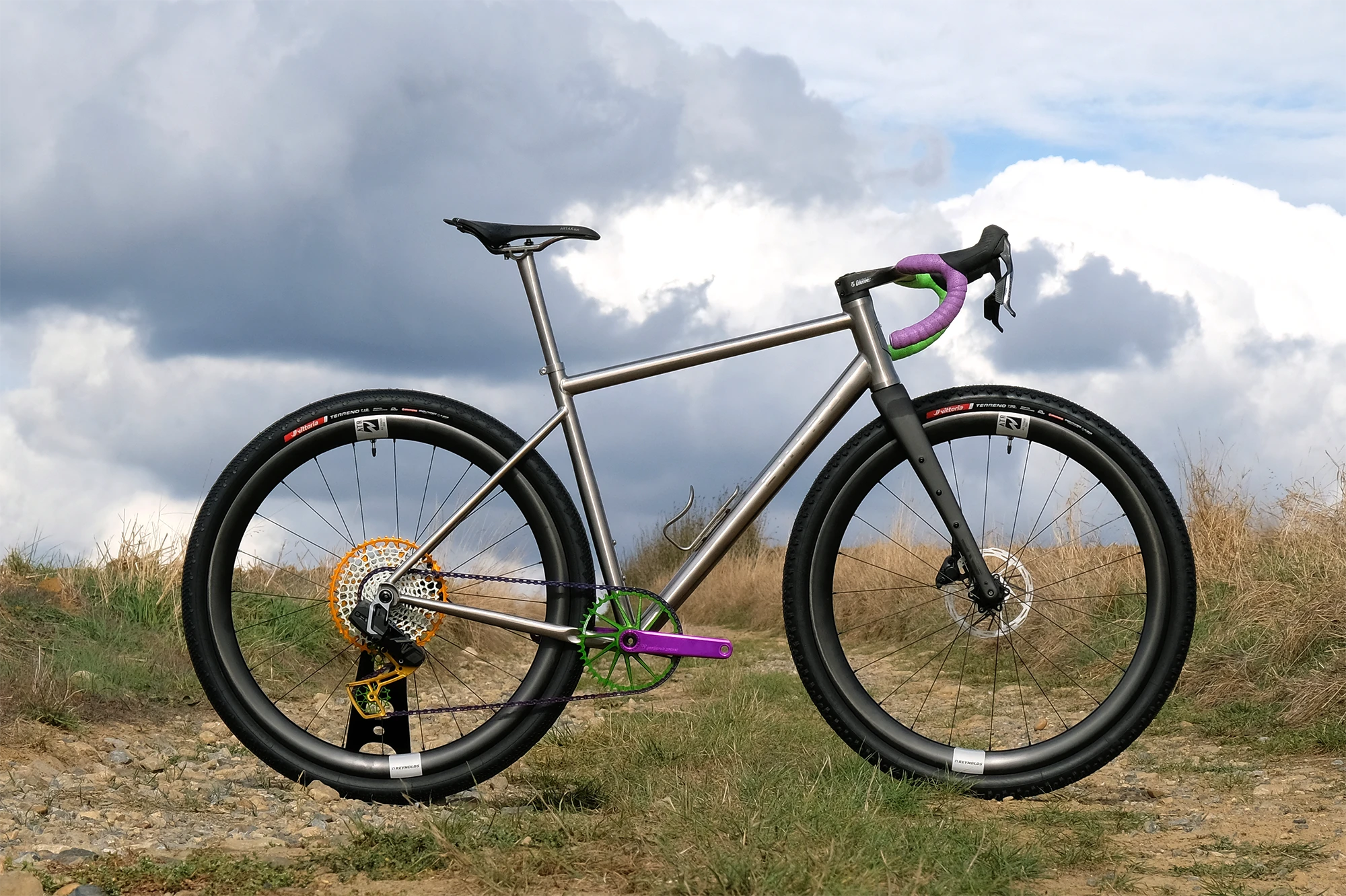 The Chiru Vagus:i titanium gravel bike in raw finish, fitted with wide tyres, photographed on a grassy track under a cloudy sky